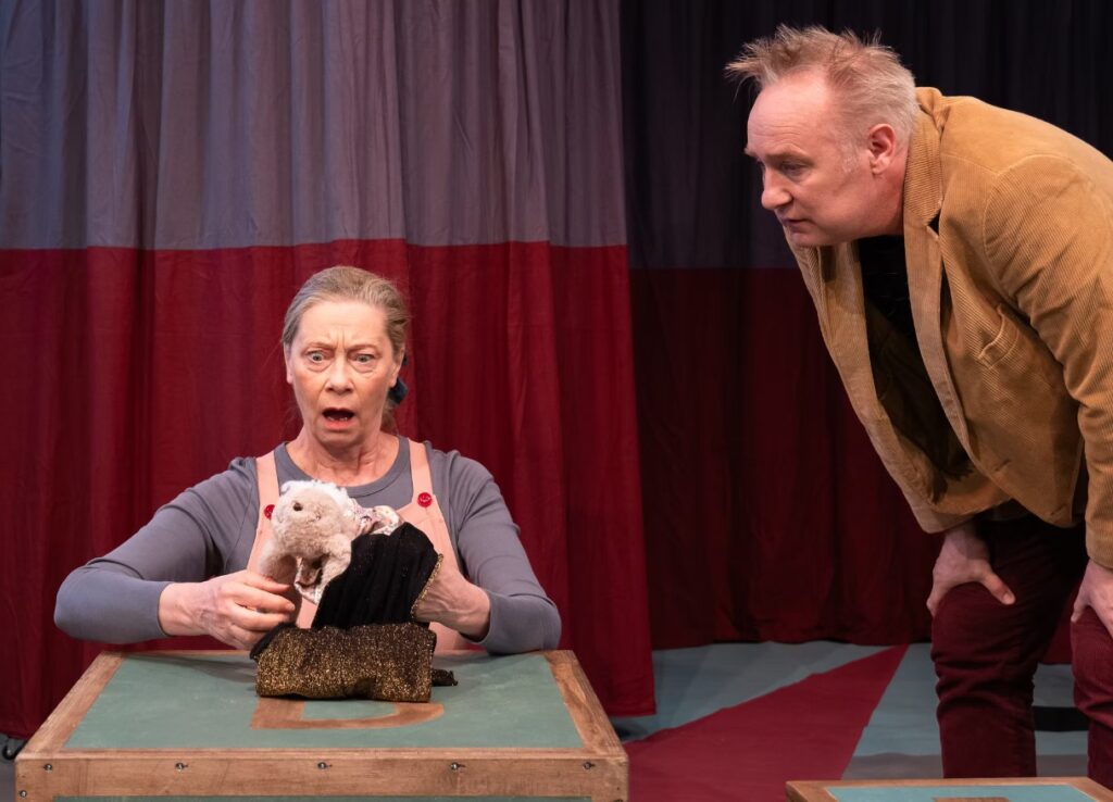 Två personer som spelar teater. En kvinna som sitter ner och håller i ett gosedjur och en man som står bredvid och tittar på.