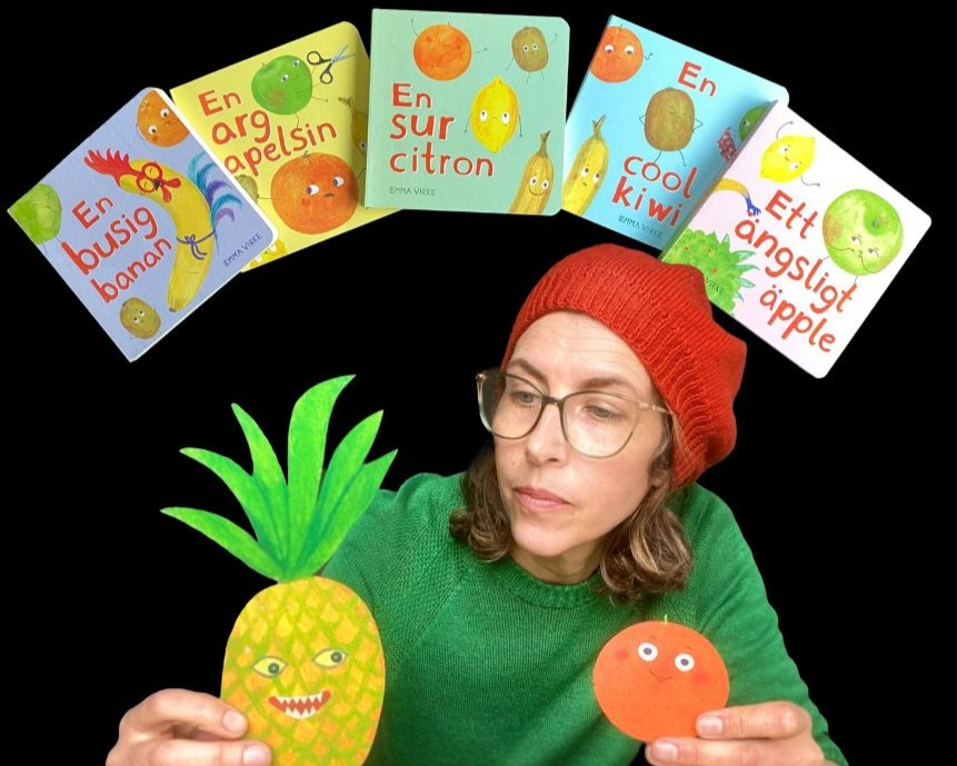 En kvinna i röd basker och grön tröja håller i en ananas och en apelsin av papper.