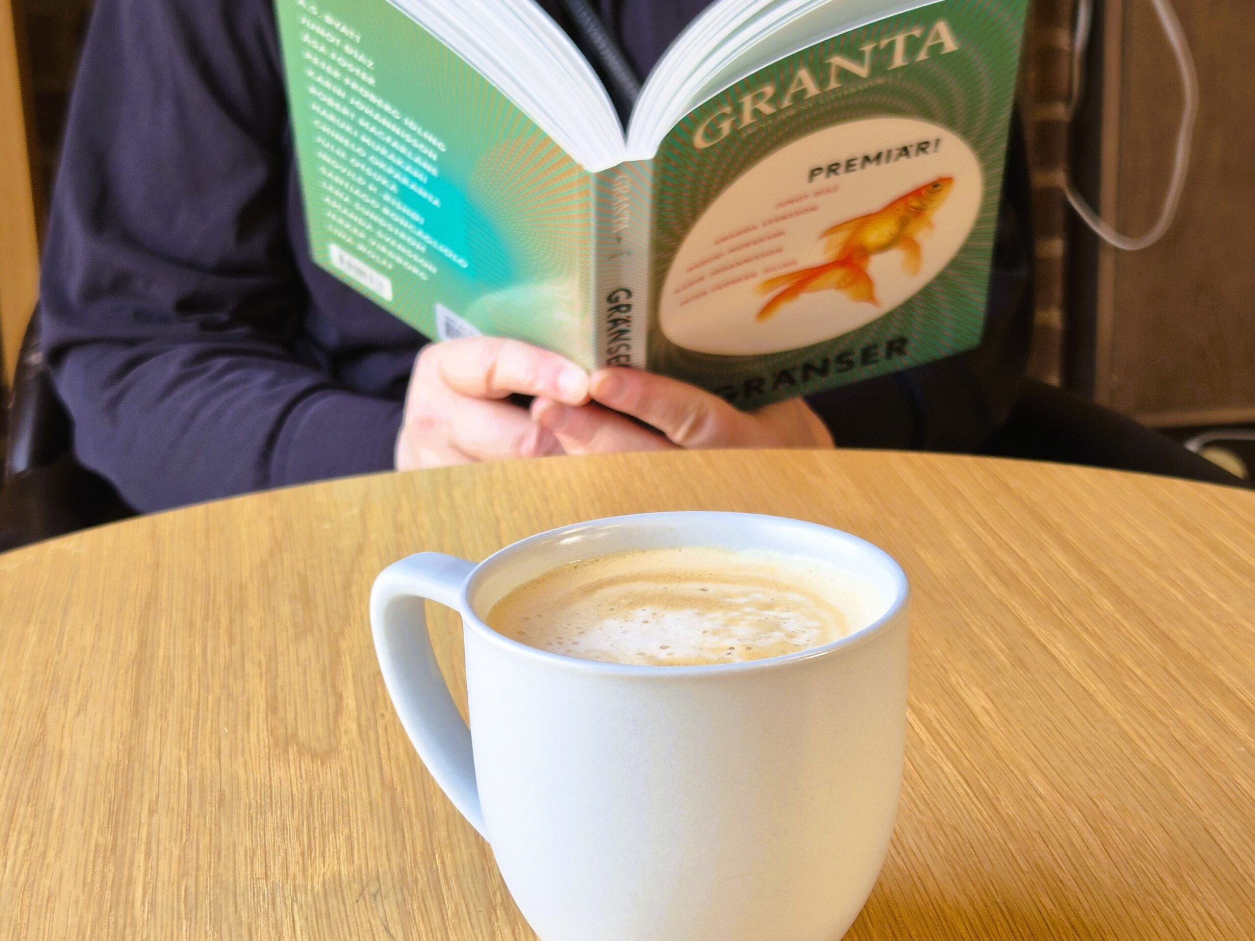 En kaffekopp står på ett bord. Bakom den syns en bok som någon håller i.