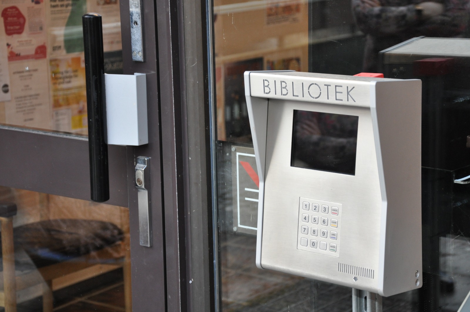 Vid sidan av en glasdörr sitter en metallpanel märkt ”BIBLIOTEK” med ett litet fönster och en knappsats.
