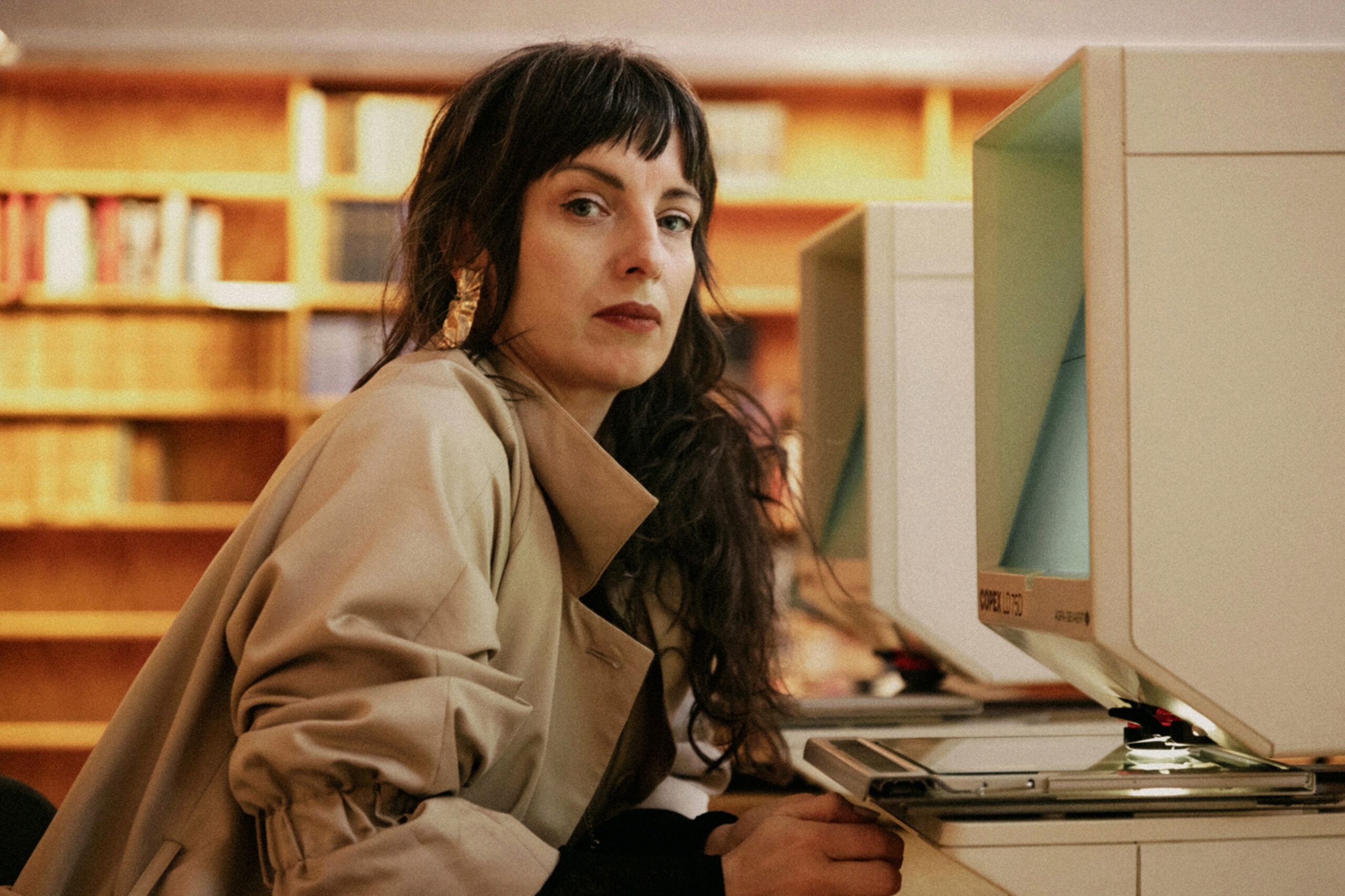 Författaren Nina Wähä sitter i ett bibliotek och tittar mot kameran. Hon har mörkt hår och en beige jacka på sig.