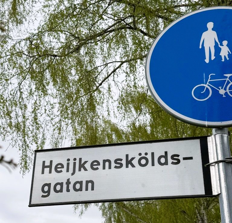Gatuskylt med namnet Heijkensköldsgatan. I bakgrunden syns en björk och skylt för övergångsställe.