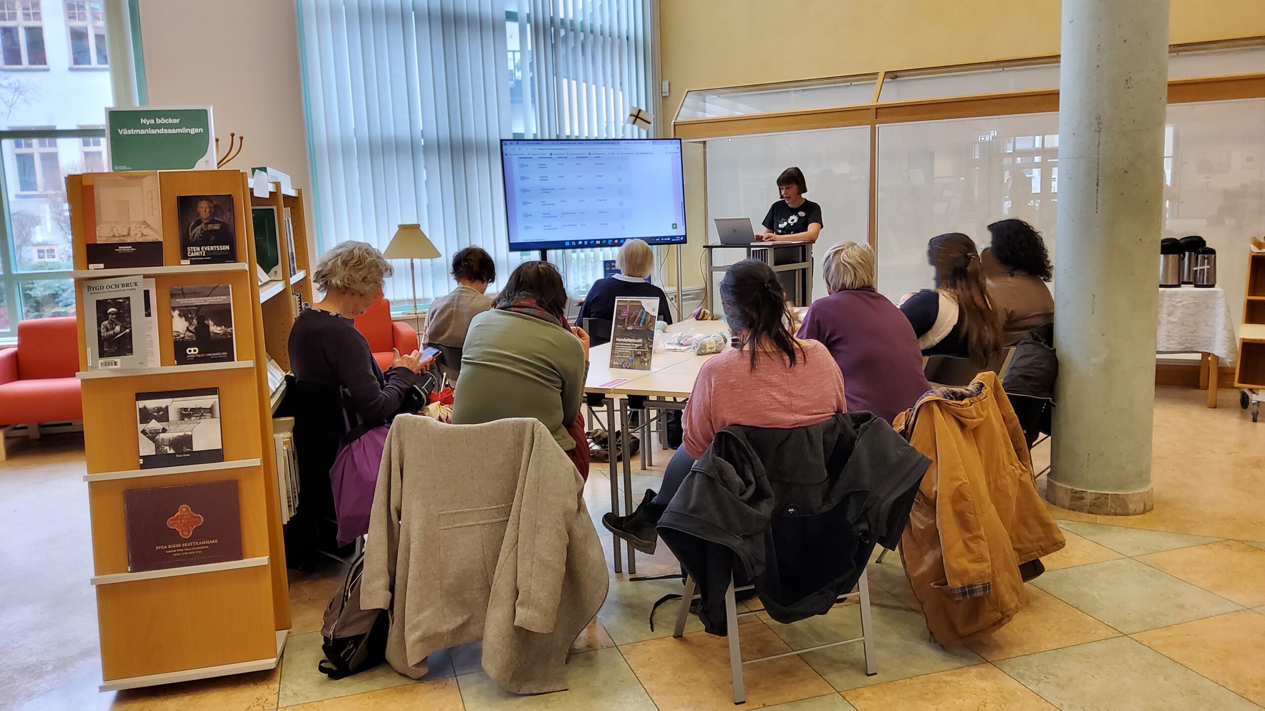 Grupp personer sitter runt ett bord på ett bibliotek och lyssnar på en ledare vid en stor skärm, med handarbetsmaterial på bordet.