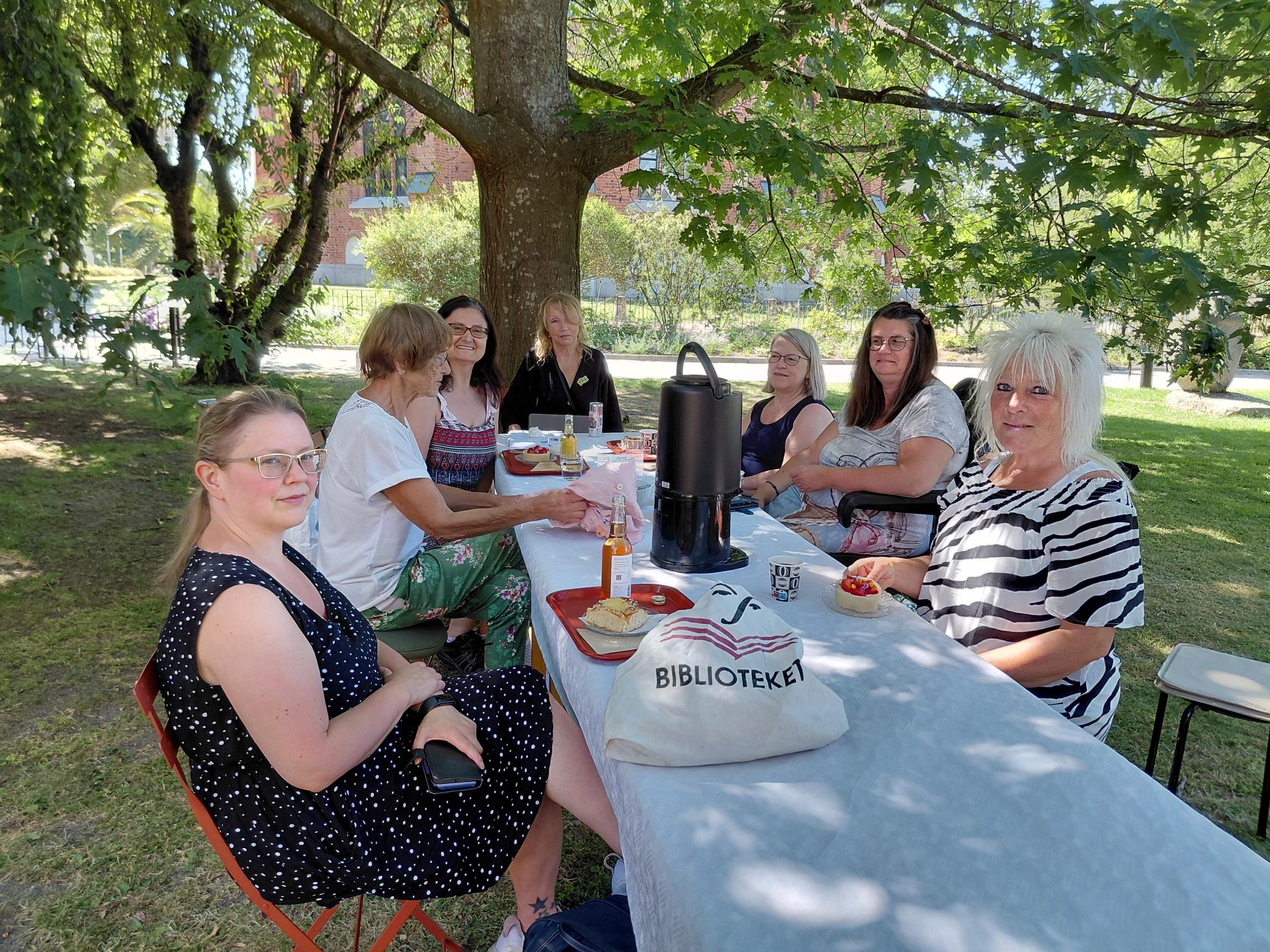 En grupp kvinnor sitter runt ett långbord utomhus under ett skuggande träd och fikar. På bordet står en kaffetermos och en tygkasse med texten ”BIBLIOTEKET”.