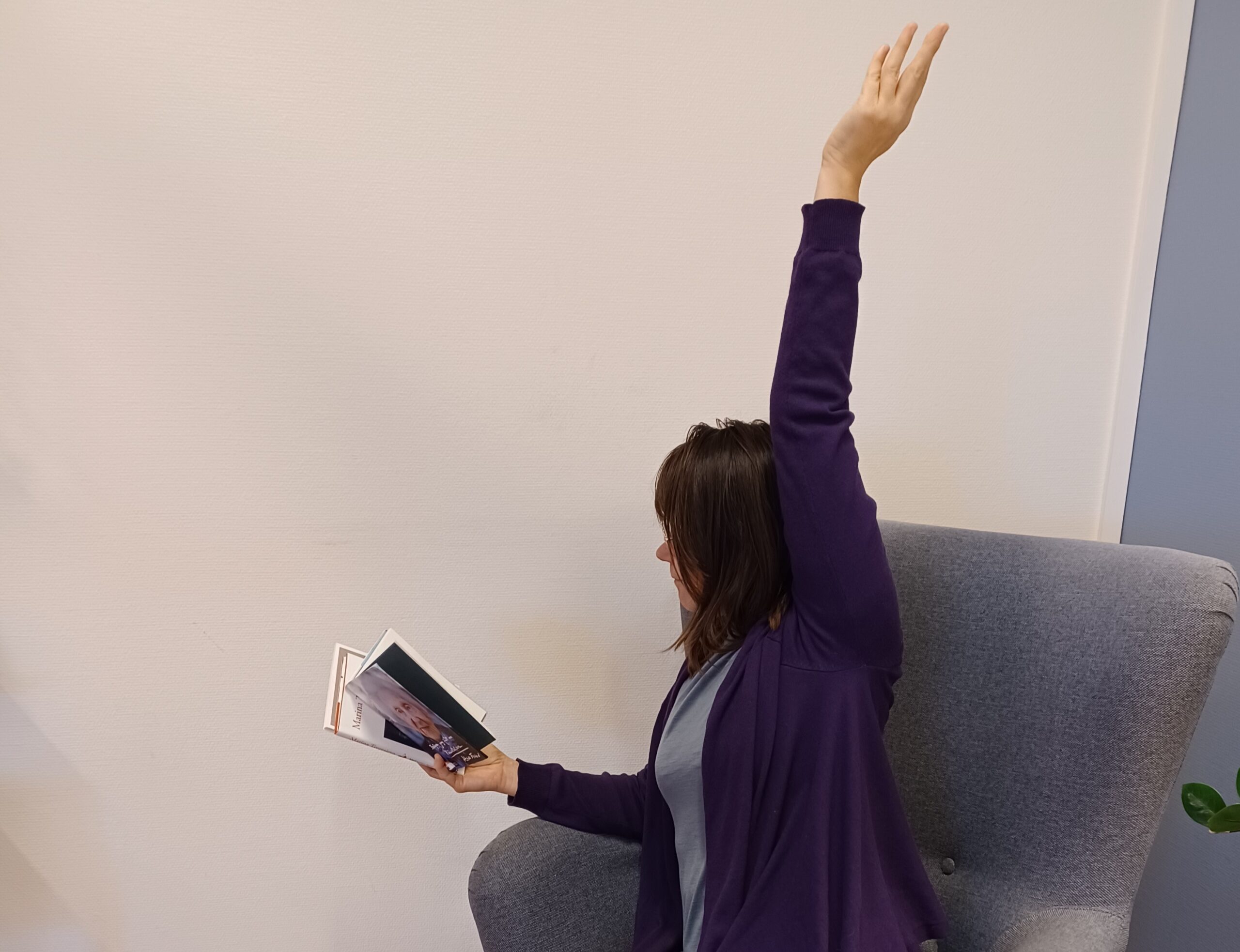 En person som sitter i en fåtölj och läser med vänster arm uppsträckt och böcker i höger hand.